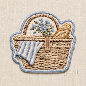 Pode incluir: Um emblema bordado em forma de cesta de piquenique com uma borda azul clara. A cesta é bege com uma alça marrom, contendo um buquê de pequenas flores azuis e uma baguete dourada. Um pano listrado azul e branco pende pela lateral da cesta.