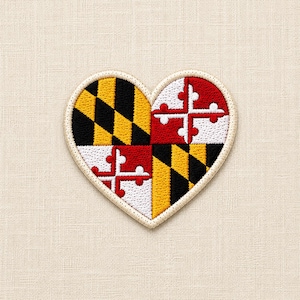 May include: A heart-shaped embroidered patch featuring the Maryland state flag design. The patch displays the iconic black and gold diagonal checkerboard pattern alongside the red and white cross bottony emblem, all bordered by a cream-colored edge.
