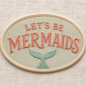 Può includere: Una toppa ricamata di forma ovale con uno sfondo verde salvia chiaro e un bordo cucito color crema. Il testo LET'S BE MERMAIDS è ricamato in rosa corallo, accompagnato da un piccolo grafico dettagliato di una coda di sirena verde acqua.