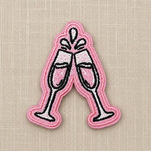May include: An embroidered patch featuring two champagne flutes clinking together in a toast. The design uses pink, white, and black thread to depict the glasses and liquid, with a pink border outlining the shape of the two glasses.