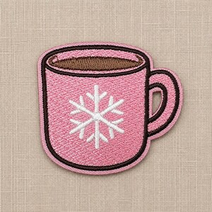 Pode incluir: Um emblema bordado cor-de-rosa em forma de caneca cheia com uma bebida castanha. A caneca apresenta um desenho de floco de neve branco no centro e é contornada com uma borda castanha escura. O bordado tem um acabamento texturizado visível.