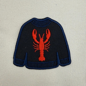 Könnte beinhalten: Ein dunkel marineblauer gestickter Aufnäher in Form eines langärmeligen Pullovers. Der Pullover zeigt mittig eine auffällige, leuchtend rote Hummergrafik. Der Aufnäher hat einen dunkelblauen gestickten Rand sowie gerippte Details an Ärmeln und Saum.