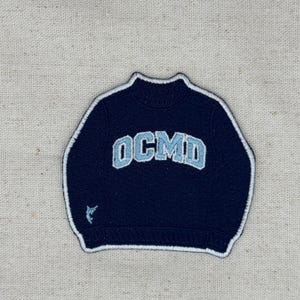 Op de afbeelding: Een donker marineblauwe geborduurde patch in de vorm van een trui met een hoge hals. In het midden staat de lichtblauwe tekst OCMD in een bloklettertype. Een klein lichtblauw visicoontje bevindt zich linksonder en de patch is wit omrand.