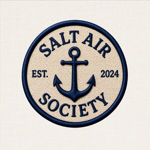 May include: A circular embroidered patch featuring a cream-coloured background with a dark blue border. A dark blue anchor is centred, surrounded by the text SALT AIR SOCIETY in dark blue lettering, with EST. on the left and 2024 on the right.