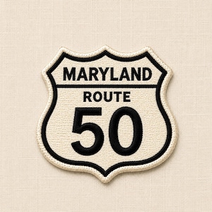 May include: A shield-shaped embroidered patch featuring a cream-coloured background with a thick black border. The text MARYLAND is embroidered in black at the top, followed by ROUTE and the number 50 in large black lettering in the centre.