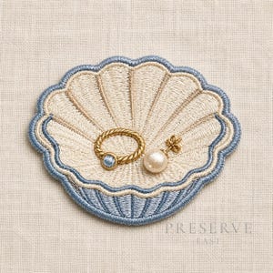 May include: An embroidered seashell-shaped tray in cream and light blue thread. Resting on the tray are a gold-toned twisted ring with a small blue centre stone and a single gold-toned earring featuring a round white pearl-like bead.