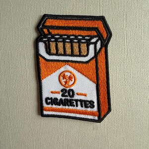 Könnte beinhalten: Ein gestickter Aufnäher in Form einer offenen Zigarettenschachtel. Das Design zeigt orangefarbene und weiße Farben mit einem schwarzen Rand. Es zeigt ein kreisförmiges Logo mit drei Sternen und den Text 20 CIGARETTES in fetter schwarzer Schrift.