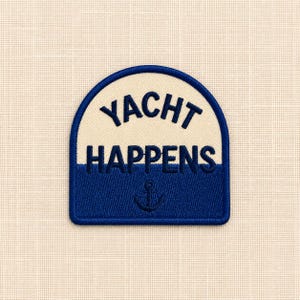 May include: An arched embroidered patch featuring a two-tone design with a cream-coloured top half and a dark blue bottom half. The text YACHT HAPPENS is stitched in dark blue, with a small dark blue anchor icon centred below the text.
