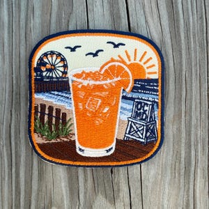 May include: A rectangular embroidered patch with rounded corners featuring a beach scene. A glass of orange drink with a slice of fruit sits in the foreground. The background shows a Ferris wheel, a lifeguard chair labeled OCMD, and birds.