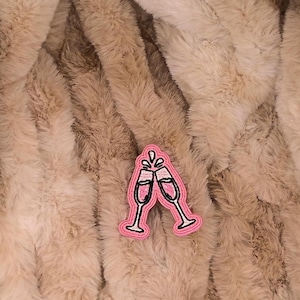 May include: A pink patch featuring two champagne glasses clinking together, with bubbles rising from the glasses. The patch has a black outline and is set against a fluffy, light brown background.
