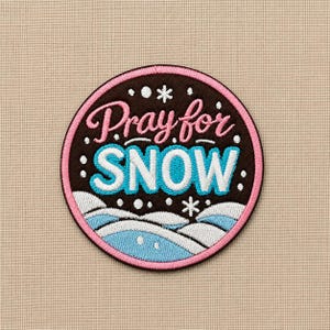 May include: A circular embroidered patch with a dark brown background and a pink border. The text reads Pray for SNOW in pink and white script and block lettering. Below the text are white and light blue embroidered snowy hills and snowflakes.