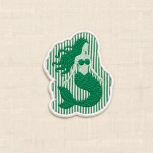 Puede incluir: Un parche bordado que muestra una sirena con cabello largo y fluido y una cola con escamas. La sirena está representada en tonos verdes sobre un fondo de rayas verticales blancas y verdes, todo ello rodeado por un borde de color blanco.