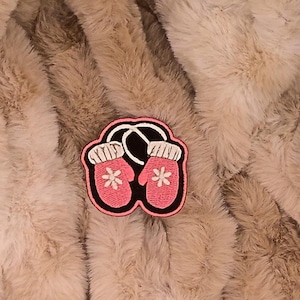 May include: A pink embroidered patch featuring a pair of mittens with white floral accents. The mittens are connected by a white string and set against a black border. The patch is on a fluffy, light brown background.