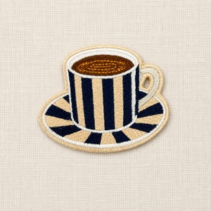 Pode incluir: Um emblema bordado apresentando uma xícara de café e pires com um padrão de listras verticais em azul marinho e creme. A xícara contém um líquido marrom com uma textura circular costurada, e a alça é de cor creme com uma borda espessa.