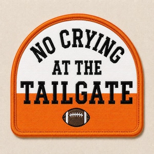 May include: A semi-circular embroidered patch featuring a white top section and an orange bottom section. The black embroidered text reads NO CRYING AT THE TAILGATE. A small brown and white football icon is centered below the text.