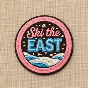 May include: A circular embroidered patch featuring a dark brown background with a pink border. The text "Ski the EAST" is stitched in pink and light blue. Below the text, white and blue snowy hills are depicted with small white snowflake accents.