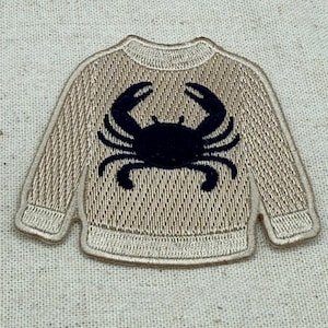 Puede incluir: Un parche bordado de color beige con forma de suéter de manga larga. El suéter presenta un gráfico de un cangrejo azul marino oscuro centrado en el pecho. El bordado muestra una textura acanalada en el cuello, los puños y el dobladillo.