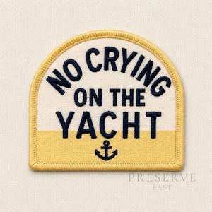 May include: A semi-circular embroidered patch featuring the dark navy blue text NO CRYING ON THE YACHT and a small anchor icon. The patch has a cream-colored upper section and a yellow lower section, finished with a yellow stitched border.
