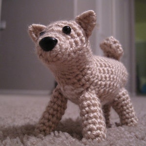 May include: A crocheted beige dog toy with black button eyes. The toy is standing on a light brown carpet.