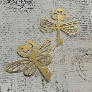 May include: Two gold-colored metal key charms with a dragonfly wing design. The keys are on a vintage newspaper background. The keys have a decorative top and a keyhole end, suitable for jewelry making or crafting projects.