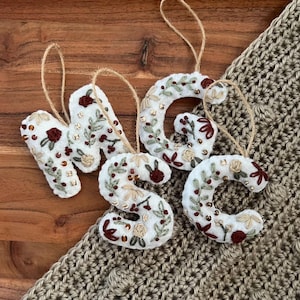 May include: Four white felt letter ornaments spelling out the initials "MGC" and "S". Each letter is decorated with embroidered floral designs in shades of red, green, and gold, with a twine loop for hanging. The ornaments are displayed on a wooden surface.