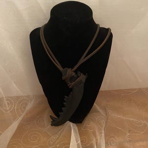 Puede incluir: Un collar con colgante negro con un diseño en forma de garra. El colgante está suspendido de un cordón de cuero marrón. El collar se exhibe en un maniquí negro.