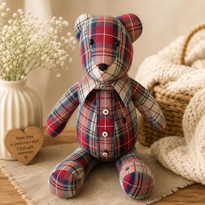 May include: A stuffed teddy bear crafted from red, blue, and white plaid fabric. The bear features a collared shirt design on its torso with four white buttons down the front, two black bead eyes, and a black nose. It has rounded ears and limbs.