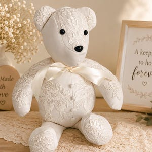 May include: A cream-colored teddy bear crafted from fabric with intricate white floral lace overlays. The bear features black bead eyes, a black nose, a satin ribbon tied around its neck, and a vertical row of small fabric-covered buttons on its chest.