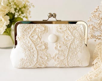 May include: A white satin clutch purse featuring an ornate cream-colored lace overlay with delicate pearl bead accents. The bag is secured with a silver-toned metal kiss-lock frame and includes a single centered white fabric-covered button detail.