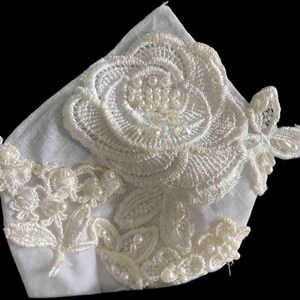 May include: White lace fabric with a large white flower design and pearl accents. The fabric is delicate and has a soft, feminine feel.
