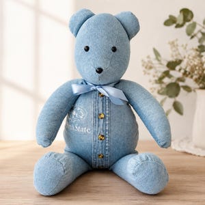May include: A stuffed teddy bear crafted from light blue denim fabric. The bear features a button-down front placket with four brown buttons, a light blue satin ribbon tied around its neck, and the embroidered text Penn State on its chest.