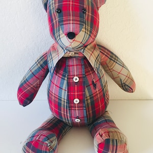 May include: A plaid teddy bear made from a repurposed shirt. The bear is sitting with its legs crossed and has a button-up shirt with a collar. The bear has black eyes and a black nose.