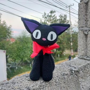 Op de afbeelding: Zwarte gehaakte knuffelkat met paarse oren en een rode strik. Het speelgoed zit op een stenen muur.