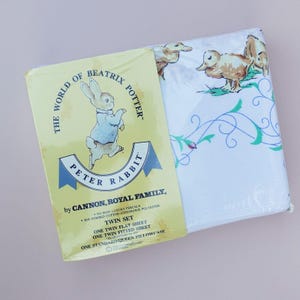 Op de afbeelding: Een twin-size beddengoedset met Peter Rabbit uit The World of Beatrix Potter. De verpakking is geel met een Peter Rabbit-illustratie. De lakens zijn wit met eenden- en bloempatronen. Inclusief een plat laken, hoeslaken en kussensloop.