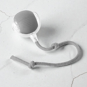 May include: A gray facial roller with a textured, rounded head, encased in a clear plastic frame. A gray rope is attached, knotted at two points. The roller is designed for skincare and facial massage.