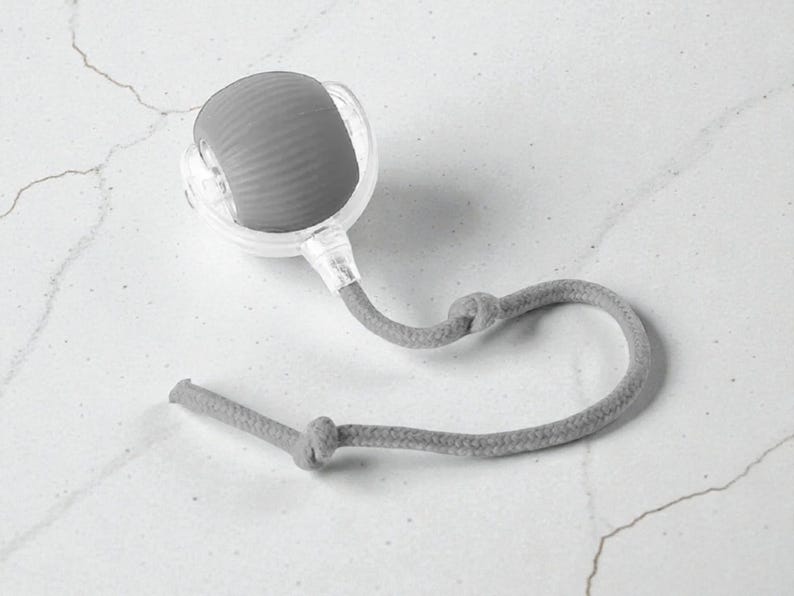 May include: A gray facial roller with a textured, rounded head, encased in a clear plastic frame. A gray rope is attached, knotted at two points. The roller is designed for skincare and facial massage.