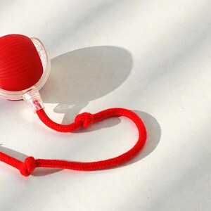 May include: A red, spherical object with a textured surface, connected to a clear plastic component and a red rope. The rope has two knots. The object casts a shadow on a white surface.