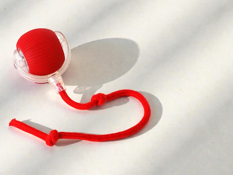 May include: A red, spherical object with a textured surface, connected to a clear plastic component and a red rope. The rope has two knots. The object casts a shadow on a white surface.