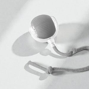 May include: A gray and clear massage tool with a textured, rounded head and a gray rope handle. The tool is designed for self-massage and relaxation. The design is simple and modern, with a focus on functionality.