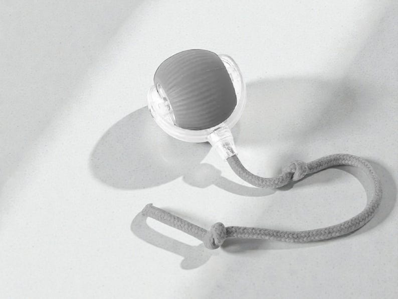 May include: A gray and clear massage tool with a textured, rounded head and a gray rope handle. The tool is designed for self-massage and relaxation. The design is simple and modern, with a focus on functionality.