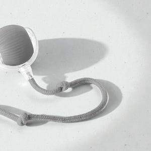 May include: A gray, handheld massage tool with a textured, rounded head and a clear plastic casing. It has a gray rope handle with a knot detail. The tool is designed for muscle relief and relaxation.