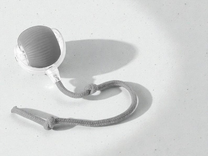 May include: A gray, handheld massage tool with a textured, rounded head and a clear plastic casing. It has a gray rope handle with a knot detail. The tool is designed for muscle relief and relaxation.