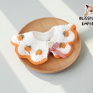 May include: A white and orange crochet pet collar with pumpkin details and a red and white checkered bow. The collar is on a wooden plate, perfect for a pet's fall wardrobe.