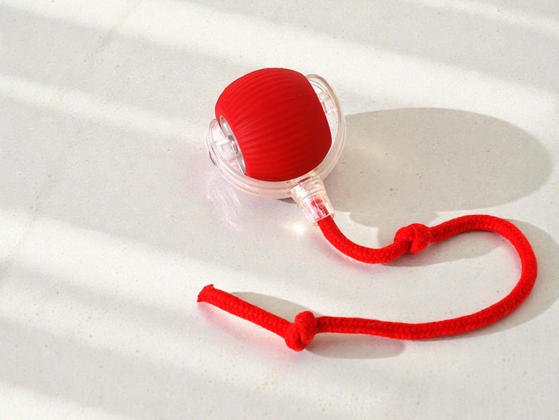 May include: A red, spherical object with a ribbed texture, encased in a clear, circular frame. A red rope is attached, knotted at two points. The object is likely a massage tool or similar wellness product.