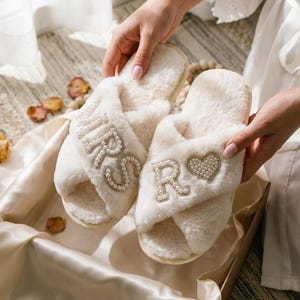 May include: A pair of cream-colored, fluffy slippers with the word "MRS" and a heart-shaped design with the letter "R" embellished with pearls. The slippers are presented in a satin-lined box.