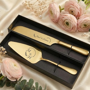 May include: A gold-colored cake knife and server set in a black box. The knife is engraved with "Mack & Hanna", and the server has "M & H" with a floral design. The set is displayed on a satin surface with pink flowers and pearls.