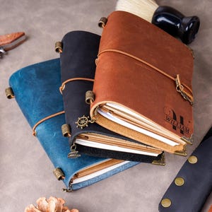 May include: Three leather journals in blue, black, and brown, with a pen, watch, and other accessories. The brown journal has the name "WILLIAM" engraved on it. The journals are closed with a leather strap.