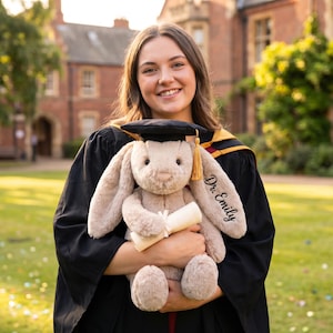 May include: A soft toy bunny wearing a graduation cap and holding a scroll. The bunny's ear is embroidered with "Dr. Emily". The person in the image is wearing a graduation gown and smiling in front of a brick building.