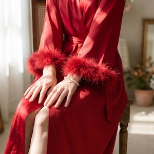 May include: A red satin robe with feather trim on the cuffs. The robe is tied at the waist and has a high slit. A pearl bracelet is visible on the wrist. The robe is worn while seated on a wooden chair.