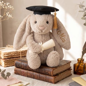 May include: A soft, light brown bunny wearing a black graduation cap with a gold tassel. The bunny holds a rolled-up scroll and sits on a stack of old books. The name "Mia" is embroidered on the ear.